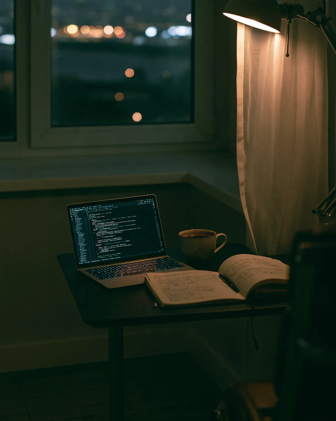 In the evening a laptop with code on a small desk by a window, beside it a cup and an open notebook, a lamp above the desk, city lights beyond the window