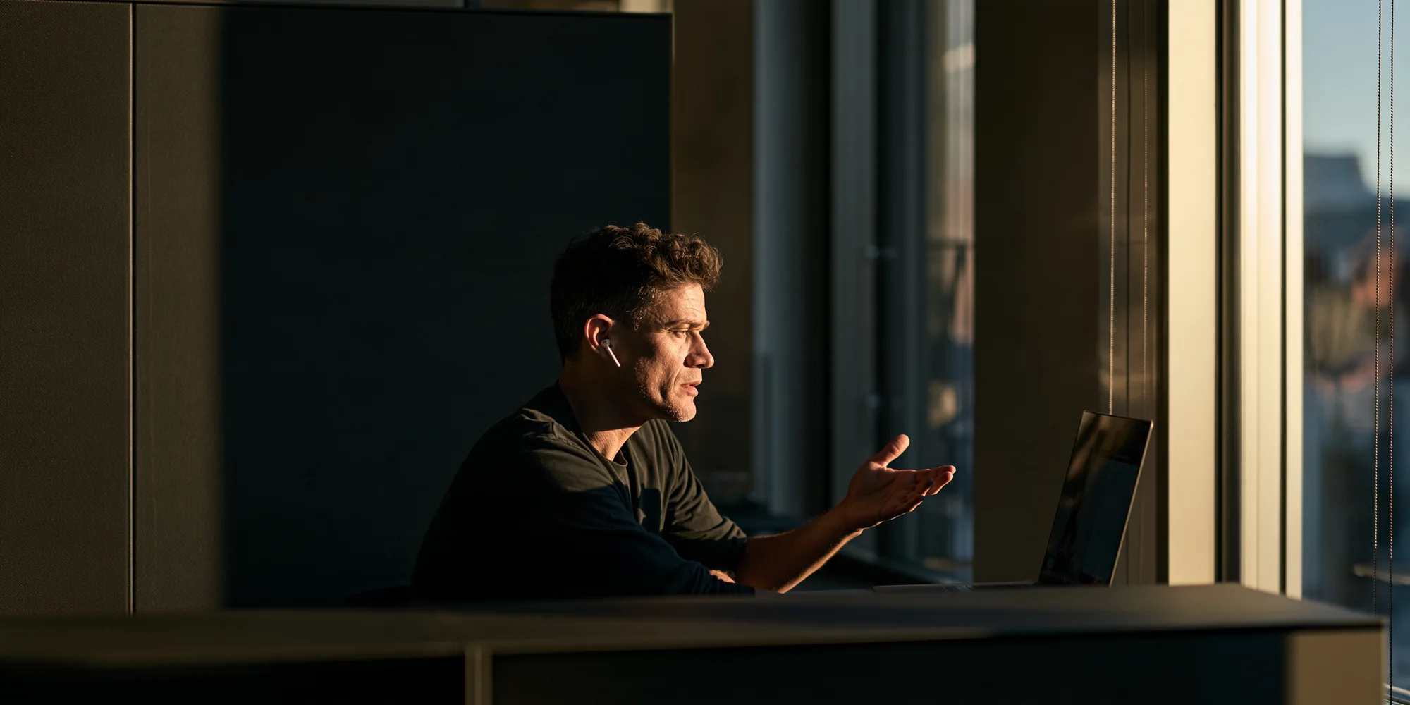 A man in a dark shirt and one earbud at a desk by a tall office window in low evening light, mid-sentence in a video conversation, one hand open, the laptop screen turned away