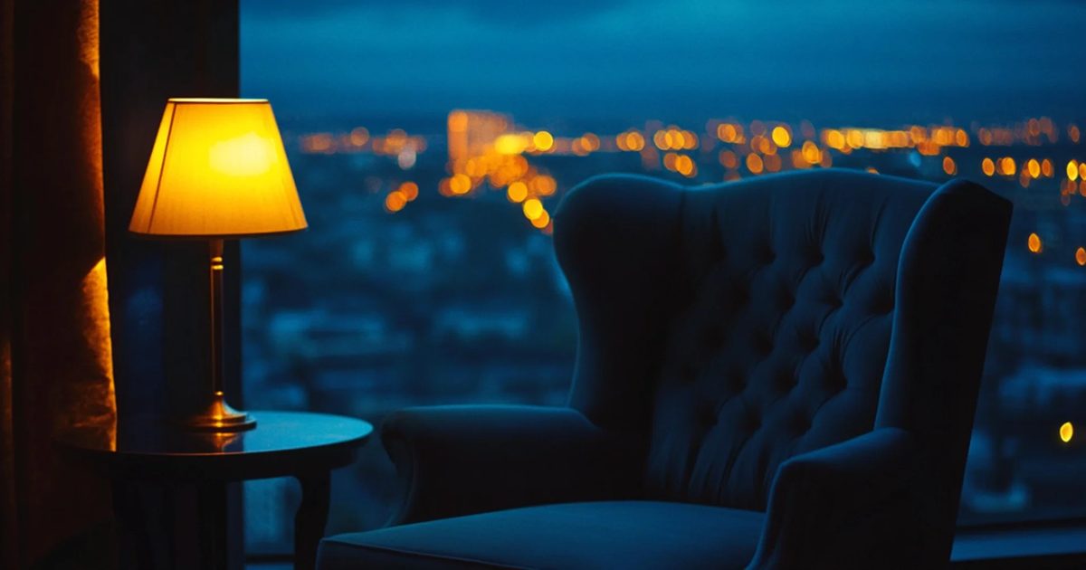 An armchair beside a lit lamp at a window, with city lights at dusk behind it.