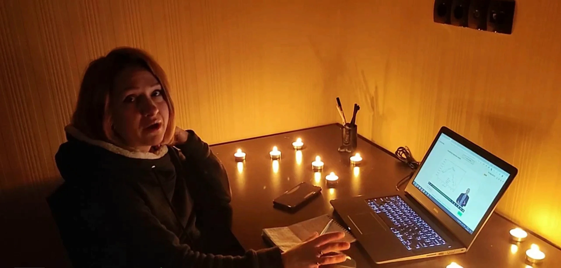 A student studying by candlelight during a power cut, her laptop open on data coursework, tealights across the table.