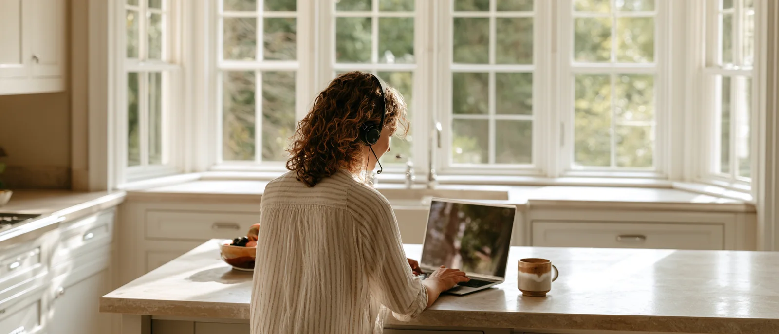 A woman with a headset on a call at a sunlit kitchen counter